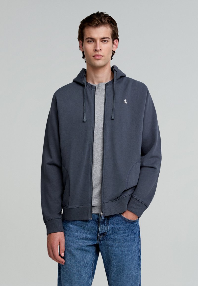 Grauer Reißverschluss-Hoodie mit Kapuze, ausgestattet mit einem kleinen Skull-Logo auf der Brust, getragen über einem grauen Shirt, kombiniert mit blauen Jeans.
