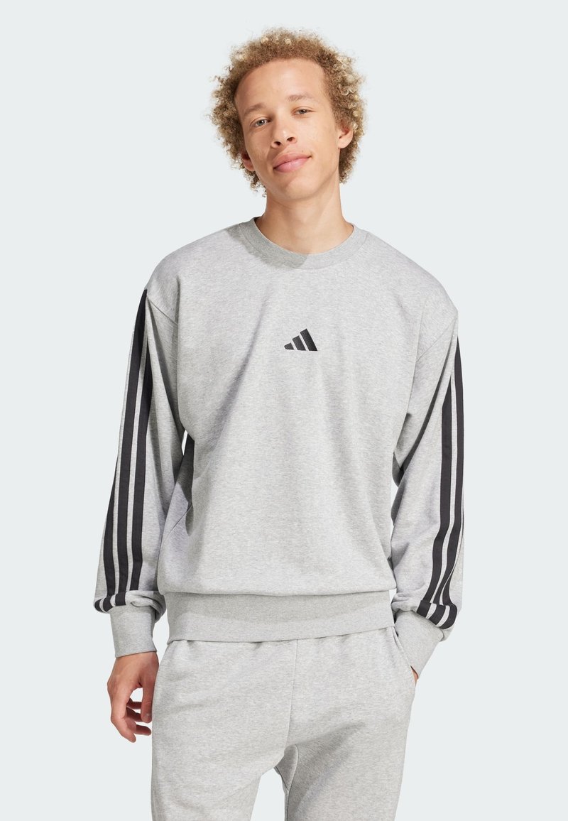 Grå sweatshirt med lange ærmer, ribbede manchetter og talje. Sorte skrå striber på ærmerne. Har et lille sort logo på brystet.