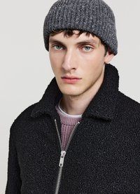 Schwarze strukturierte Jacke mit Reißverschluss und Kragen, darüber ein hellrosa geripptes Sweatshirt getragen. Graue gestrickte Mütze. Neutraler Hintergrund.