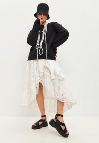 Černá oversized mikina, bílá krajková maxi sukně s vrstveným lemem, černé chunky sandály s širokými pásky a tkaný černý klobouk bucket.