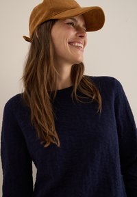 Pull en motif marine avec une finition texturée, associé à une casquette de baseball en daim beige, affichant un profil décontracté sur un fond neutre.