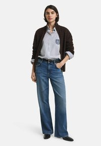 Vrouw in een bruine kabelgebreide zip-up jas, een blauw gestreept overhemd met insignia, blauwe wijde jeans en zwarte puntige schoenen.