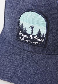 Marineblå caps med teksturert stoff, med et rundt brodert merke av en golfer blant trær. Teksten sier "Stripes & Pines National Open."
