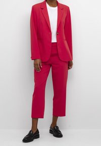 Felroze pak bestaande uit een op maat gemaakte blazer en korte broek, gecombineerd met een wit T-shirt en zwarte loafers. Gladde stoftextuur.