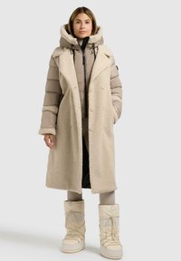 Beige schapenvachtjas gelaagd over een pufferjack, met een grote capuchon, dubbele knoopsluiting aan de voorkant en hoge witte laarzen met veters.