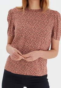 Blouse à manches courtes en rose poudré avec un motif tacheté noir et crème éparpillé, dotée d'un col rond et d'un tissu doux et texturé.