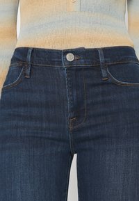 Jeans de mezclilla de alta cintura en azul oscuro con cierre de botón, que presentan un sutil contraste de costuras y un diseño de cinco bolsillos.