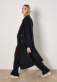 Langer schwarzer Mantel mit doppelt knöpfbarem Verschluss, texturiertem Stoff und taillierter Form, kombiniert mit dunklen Hosen und hellen Schuhen.