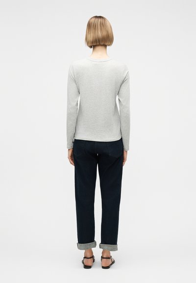 Γυναίκα με κοντά ξανθά μαλλιά στέκεται με την πλάτη, φορώντας ανοιχτό γκρι μπλουζάκι με μακριά μανίκια, σκούρα τζιν με γυρισμένα μανίκια και μαύρα σανδάλια.