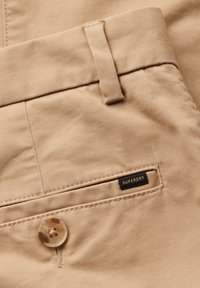 Pantaloni in cotone beige con una texture liscia, chiusura con bottone e un'etichetta nera con la scritta "SUPERDRY" sulla tasca.