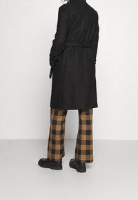 Sort trenchcoat med bælte i taljen, parret med brune og sorte ternede bukser med vidde og sorte klodsede støvler. Glat tekstur og skræddersyet pasform.