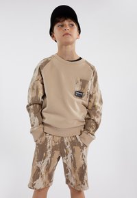 Felpa beige a girocollo con maniche a motivo marrone, dotata di tasca frontale e patch con logo, abbinata a pantaloni corti con lo stesso motivo.