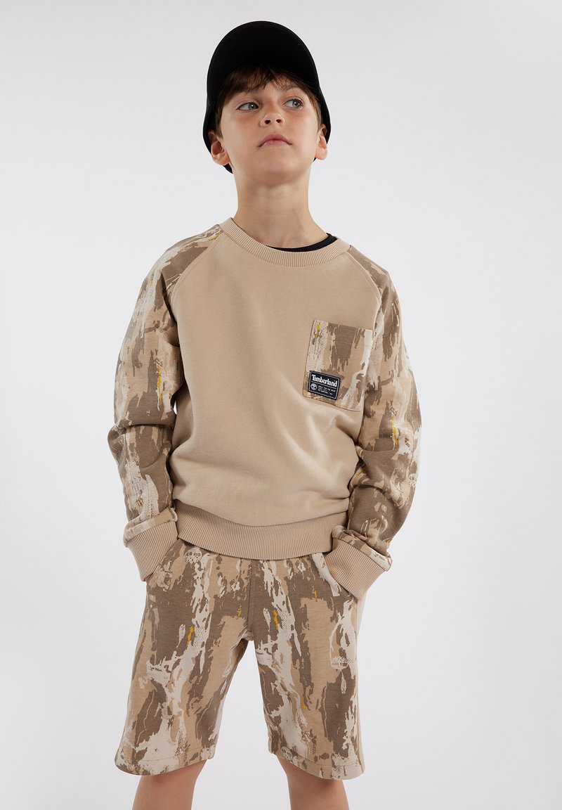 Felpa beige a girocollo con maniche a motivo marrone, dotata di tasca frontale e patch con logo, abbinata a pantaloni corti con lo stesso motivo.