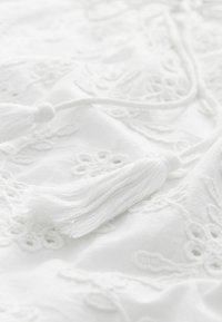 Tissu blanc présentant des motifs floraux brodés et des pompons. La texture est douce, avec des détails complexes et une couleur homogène.
