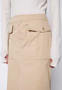 Jupe beige à taille haute avec grandes poches à rabat boutonnées portée avec un haut blanc à manches longues en maille côtelée avec des ouvertures pour les pouces.