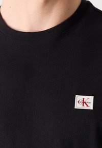 Czarna bawełniana koszulka z krótkim rękawem z okrągłym dekoltem, z małą łatką logo Calvin Klein w białym i czerwonym kolorze na piersi.