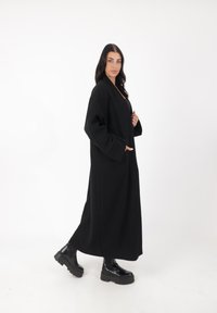 Langer schwarzer Mantel aus dickem Stoff, mit lockerer Passform, Seitentaschen und minimalistischem Design, kombiniert mit schwarzen Plateauboots.