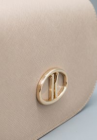 Borsa in pelle beige texturizzata con una fibbia ovale dorata lucida con due barre verticali su una superficie azzurro chiaro.