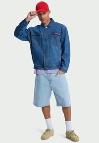 Denim jacket in faded blue with buttoned front, two chest pockets, and logo. Paired with light blue shorts, white socks, and beige shoes.