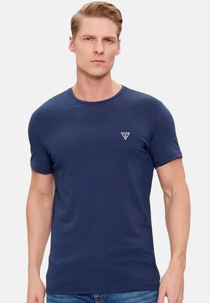 Homme aux cheveux blonds courts portant un t-shirt bleu marine ajusté à col rond avec un petit logo sur la poitrine, posé devant un fond clair uni.