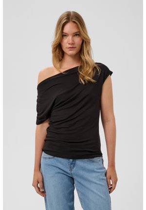 Jeune femme aux longs cheveux blonds porte un haut noir à une épaule et un jean bleu clair, debout devant un fond gris clair uni.