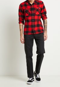 Camisa de franela a cuadros en rojo y negro con bolsillos con botones, combinada con pantalones negros a medida y zapatillas negras con suelas blancas.