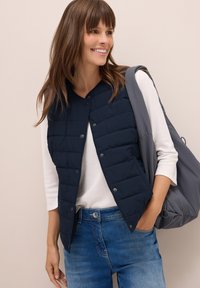 Gilet puffer blu marina con design trapuntato, dotato di scollatura rotonda e bottoni a pressione. Indossato sopra una maglietta a maniche lunghe bianca e jeans blu.