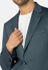 Blauer Blazer aus strukturiertem Stoff, mit einem einzelnen Knopf, Schalkragen und einem kontrastierenden Innenfutter. Darunter ein weißes T-Shirt.