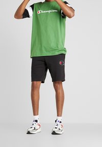 Camiseta de algodón verde con mangas blancas y negras, con el logo de "Champion". Se lleva con pantalones cortos negros que muestran un logo bordado y zapatillas deportivas.