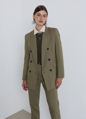 Frau mit kurzen dunklen Haaren, die einen olivgrünen, doppelt geknöpften Blazer und passende Hose über einem schwarzen Top und einem weißen Hemd mit Kragen trägt.