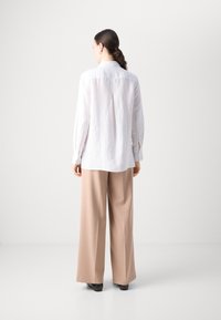 Camicia bianca in lino con orlo curvo, polsini abbottonati e piega sul retro, abbinata a pantaloni larghi beige e scarpe nere.