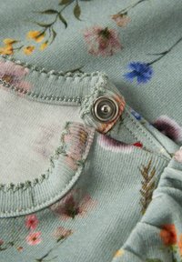 Bloemstof met een lichtblauwe achtergrond, voorzien van een ronde halslijn en een zilveren drukknoopsluiting. Zachte textuur met gerimpelde randen.