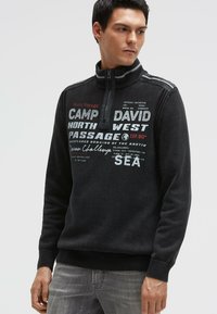 Schwarzer Zip-up-Sweatshirt mit strukturierter Strickoptik, versehen mit weißen und roten Textdruck-Designs. Gerippte Bündchen und Saum, hoher Kragen.
