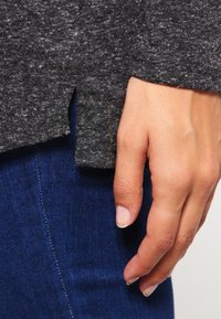 Grau strukturierter Pullover mit seitlichem Schlitz, kombiniert mit dunkelblauen Jeans. Hand ruht auf dem Saum des Pullovers.