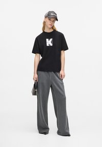 Juodas per didelis marškinėliai su grafiniu 'K' spaudiniu; pilkos plačios kojų sportinės kelnės; pilkas beisbolo kepuraitė; maža juoda rankinė su dirželiu.