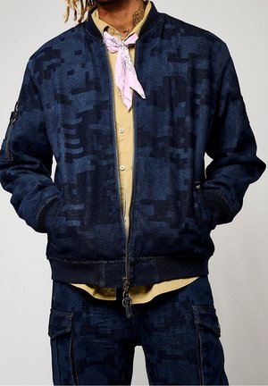 Homme portant une veste et un pantalon camouflage numérique bleu foncé avec une chemise beige et un foulard rose clair, les mains dans les poches de la veste.