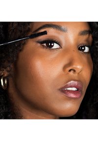 Brosse de mascara noire tenue près de l'œil d'une femme. Sa peau a une teinte chaude, ses lèvres brillantes sont d'un rose doux. Des cheveux bouclés encadrent son visage. Une boucle d'oreille en or est visible.
