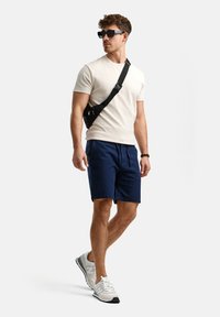 Light beige t-shirt, navy shorts, black crossbody bag, and white sports shoes with gray accents, paired with dark sunglasses.