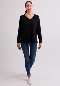Czarny sweter w serek z miękkiego materiału, z długimi rękawami i luźnym krojem, w połączeniu z niebieskimi skinny jeansami i białymi trampkami.