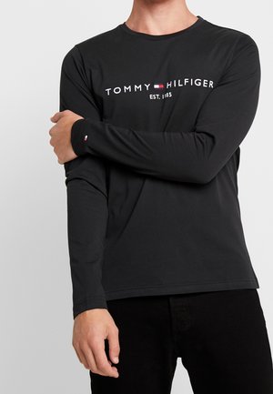 Homme portant un t-shirt à manches longues noir Tommy Hilfiger avec logo et texte "Est. 1985" sur la poitrine, croisant un bras sur l'autre.