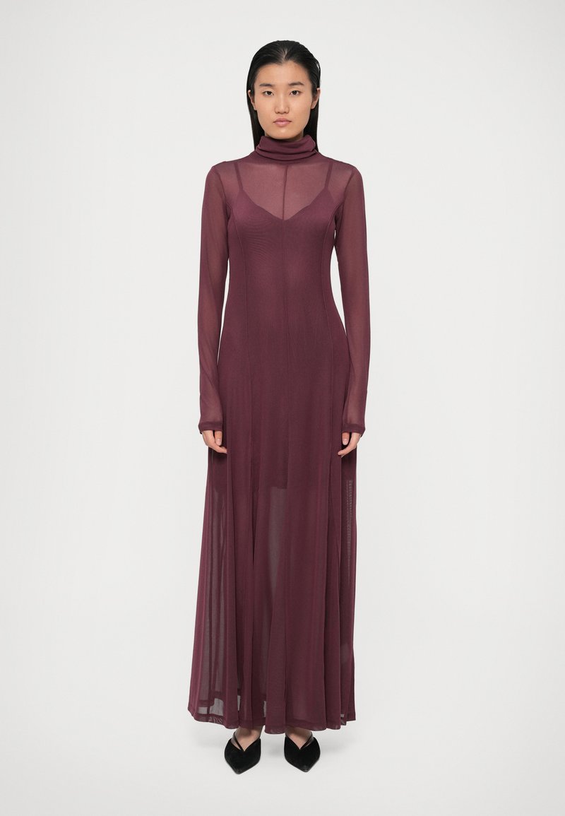 Abito lungo bordeaux a maniche lunghe con scollatura alta e tessuto trasparente, caratterizzato da un corpetto aderente e una gonna fluida. Scarpe nere a punta visibili.