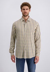 Mann in einem langärmligen gelb, braun und weißen karrierten Hemd mit Knöpfen und blauen Jeans, der vor einem schlichten Hintergrund steht.