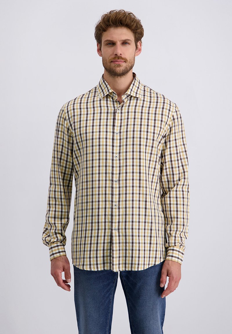 Mann in einem langärmligen gelb, braun und weißen karrierten Hemd mit Knöpfen und blauen Jeans, der vor einem schlichten Hintergrund steht.