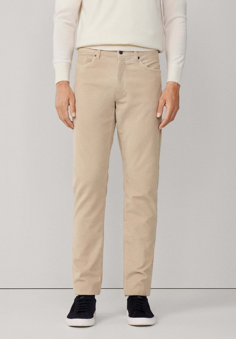 Uomo che indossa pantaloni beige a gamba dritta, maglione bianco e scarpe da ginnastica nere, in piedi contro uno sfondo grigio chiaro uniforme.