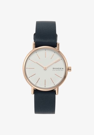Cadran rond blanc avec des marqueurs et aiguilles en or rose, bracelet en cuir noir, et logo SKAGEN Denmark sur le côté droit.