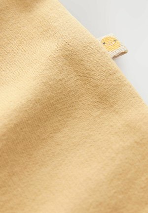 Baumwollstoff in sanftem Gelbton mit glatter Textur. Verfügt über ein kleines Etikett mit einem lächelnden Sonnensymbol.