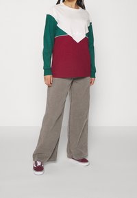 Långärmad skjorta med färgblockering i vitt, grönt och vinrött med en triangeldesign och vit logotyp; i kombination med vida ljusgrå manchesterbyxor.