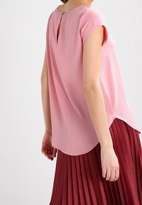 Blouse rose clair à manches courtes, avec un détail d'ouverture en forme de clef au dos et une coupe décontractée, associée à une jupe plissée bordeaux. Texture lisse.