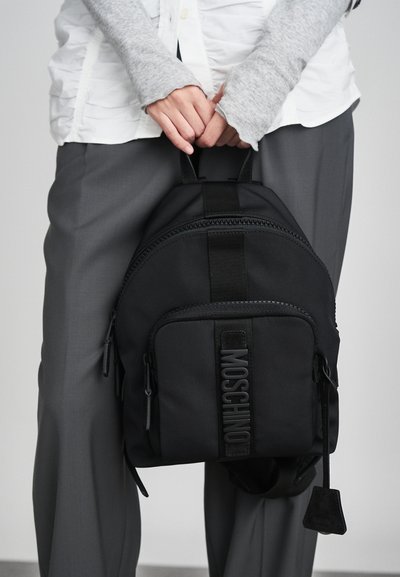 Άτομο με γκρι παντελόνι και λευκό πουκάμισο κρατάει μια μαύρη τσάντα Moschino από τις πάνω λαβές μπροστά του.