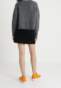 Femme portant un pull gris, une jupe noire et des baskets orange vif, se tenant sur un sol blanc uni devant un fond blanc.